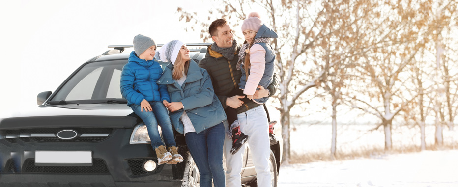 A happy family of four, dressed in winter hats and jackets, poses outside next to a black SUV in a snow-covered landscape.