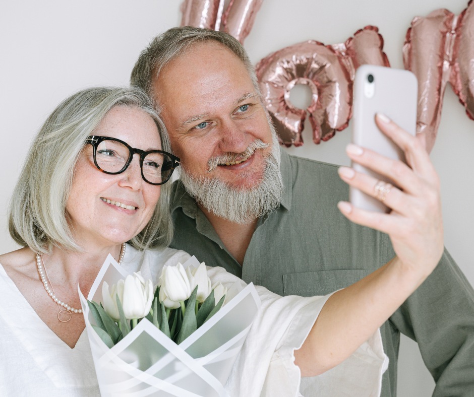 A smiling mature couple takes a selfie, with the woman holding a bouquet of white tulips and rose gold balloons in the background