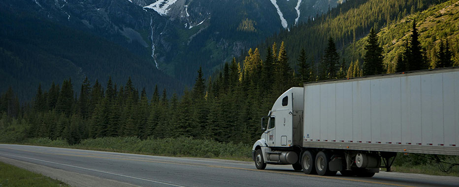 White semi-truck driving on a highway surrounded by dense pine forest - https://www.jjinsure.com/
