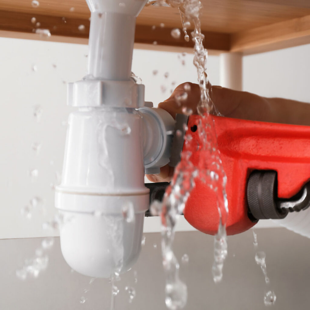A white plastic sink drain pipe under a wooden countertop with water spraying and dripping from a leak.