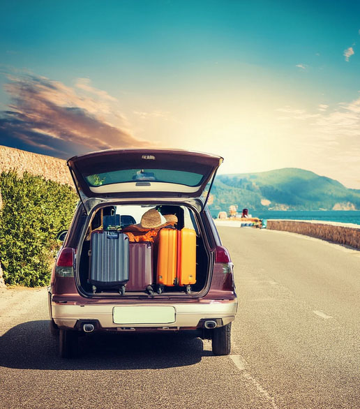 A car with an open trunk filled with colorful luggage and a straw hat is parked on a coastal road
