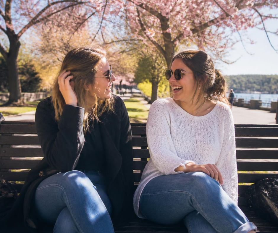 Two women wearing sunglasses smiling and talking while sitting on a park bench near a waterfront.